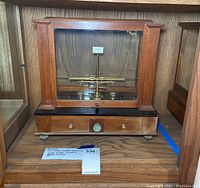 Front view of oak and glass display case enclosing brass beam scale on black slate base with front drawer.