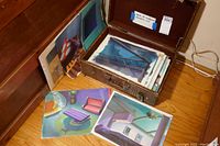Opened vintage case showing multiple stacked wrapped background animation cels with a few laid out in front showing interior scenes and architectural backgrounds.