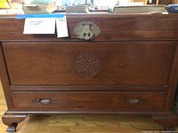 Front view of wooden cedar chest showing carved circular design, metal clasp, and bottom drawer with handles.