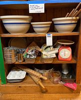 Shelf showing four Roseville Ohio ceramic mixing bowls with blue stripe design, along with vintage baking tools including cupcake pans, pans, sifters, cutters, scale, rolling pins, spoons, and measuring cup.
