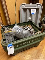 Green Rubbermaid ActionPacker container lid open filled with mixed metal climbing hardware and black webbing or fabric, labeled 'Climbing Hardware' with paper tag.