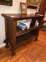 Side angle view of the solid wood console table showing the top surface and middle adjustable shelf with slatted design, placed in a room with wooden flooring and wallpaper.