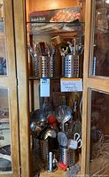 Photo showing multiple stainless steel cooking utensils in perforated metal holders inside a wooden cabinet