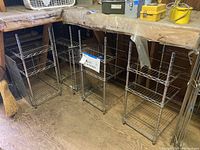 Four chrome-plated metal wire shelves standing in a row under a workshop bench, each with 3 tiers and cylindrical support poles. Shelves show surface dirt and need cleaning.