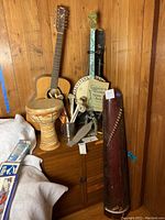 Photo showing Yamaha guitar, tenor banjo with Guckerts chords booklet, djembe drum with ceramic base, assorted drumsticks, and damaged Koto harp leaning against the wall.