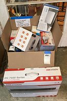 Cardboard box containing several electronics boxes: Hikvision NVR, Lefun camera, Foscam camera, Lutron dimmer kit, Arlo router box, small voice recorder box