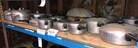 Shelf of multiple aluminum cookware pieces with lids including saucepans and drop roaster
