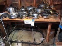 Photo showing full set of 13 pots and pans arranged on wooden surface above wrought iron rack under table.