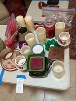 Overview of assorted candles and candle holders arranged on white folding table, showing variety of items in lot including pillar candles, jar candle, flameless candles, and floral ceramic holders.