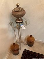 Overview showing carved wood jar on top of glass-top metal side table, with two dark amber glass jugs placed nearby on floor.