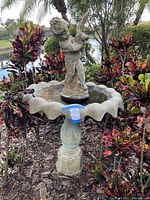 Front and angled view of concrete bird bath with scalloped edge basin and boy holding duck statue in center, showing overall condition and placement in garden setting.
