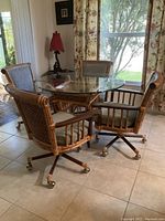 Photo of the round glass top dining table and four rattan rolling chairs arranged around it in a tiled kitchen space with windows and curtains.