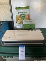 FoodSaver vacuum sealer machine on green surface with owner's manual and box of vacuum sealing rolls behind it.