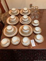 Six fully stacked place settings of white Limoges china with gold borders and six water glasses arranged on wooden table.