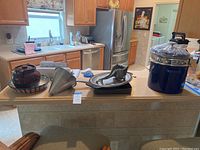 Full view of lot items on kitchen counter including stoneware crock, vintage aluminum citrus press, stove top griddle, ceramic dish, and Cuisinart stockpot