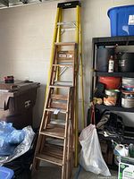 Photo showing the three ladders stored in garage: yellow 6 ft Werner fiberglass ladder at back, vintage 4 ft wooden step ladder in front, and taller 8 ft yellow Husky fiberglass ladder on the side.
