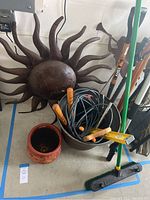 Photo of a large decorative metal sun garden wall art, terra cotta planter, garden tools including rake, broom, and a bundle of cables or hose in a metal bucket.