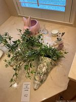 Overall display of vintage bathroom decor on countertop near window, including pink ceramic bunny planter, bone china wall vases holding faux green ivy, small clear glass perfume bottle, and various small ceramic and porcelain pieces.