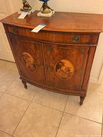 Front view of the vintage wooden console table showing curved front, decorative inlay depicting classical scenes, drawer, and cabinet doors with metal handles.