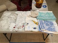 Table showing a variety of lace doilies, embroidered linens, and folded vintage tablecloths in different patterns and colors laid out on a table.