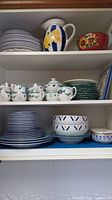 Shelf with assorted dishware including white plates, a yellow floral water jug, sunflower patterned bowl, and floral printed teapots and cups.