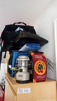 Photo showing stacked camping gear on wooden cabinet including boots, folded seating pads, blue cooler bag, battery lantern, and box of smoking chips.