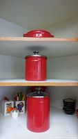 Three red ceramic canisters of varying sizes with silver lids stacked on shelves