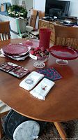 Full view of lot items arranged on wooden table, showing red glass chargers, centerpiece charger, cake plate, napkin plates, decorative bowl with artificial fruit and linens.