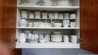 Photo of upper cupboard filled with white ceramic dishes including ramekins, bowls, cups, plates, and sugar/creamer sets.