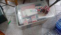 Clear plastic storage box with card decks, scorepads, and game materials on tiled floor.