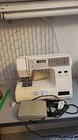 White Janome Memory Craft 7000 electronic sewing machine on a table with foot pedal and power cord visible