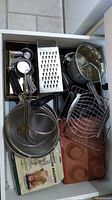 Top view of kitchen drawer holding various kitchen accessories including graters, sieves, measuring cups, roasting pans, and a muffin tray.