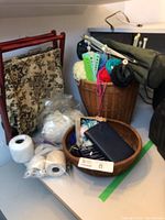 Photo of woven basket containing yarn, knitting tools, flower cushions, white yarn balls and string spool, and brown bowl with small tools.