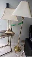 Cream colored table lamp with three candle-style bulbs, brass base, and fabric shade next to brass floor lamp with matching fabric shade.