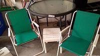View of overall lot showing round glass patio table, two green folding chairs, and small white plastic table.