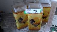 Five glass canisters arranged on a countertop, showing colorful rooster and sunflower prints with lids on.