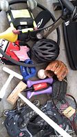 Curling brooms, baseball gloves, baseball, rollerblades, bike helmet, bike seat, hand weights, tennis racket, delivery balance bar, and cycling shirt arranged on floor.