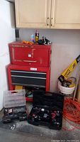 Photo of two stacking tool boxes, one red and one black with red details. Items on and around boxes include spray can, gloves, and saw in bucket.