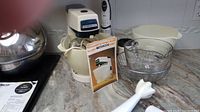 Kenwood Chef mixer with two bowls, various attachments including beaters and whip, Pyrex glass bowl, and manual on countertop.