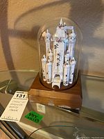 Castle made of rolled recycled Disney paper towers inside a glass dome on a wooden base with label residue and stains visible on the wood.