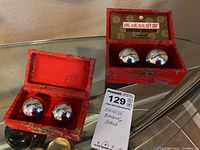Two sets of polished metallic Chinese Baoding balls in red fabric-covered boxes on a glass surface with a MaxSold lot card