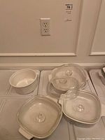 Five pieces of Corning Ware including 2 square casseroles and 2 glass lids plus one small round white dish without lid, showing scratches and chips.