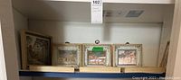 Seven framed pictures with gold tone frames arranged on shelves, showing various landscape and decorative artworks.