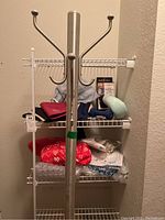 Full view of silver metal coat rack with four double hooks and wire shelving unit behind it containing various items including hand weights, heating pad, neck pillow, and apparel.