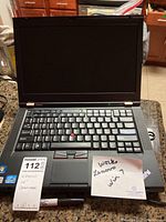 Front view of Lenovo ThinkPad T420 laptop open showing keyboard, TrackPoint, and display with 'Works Lenovo Win 7' note and tag.