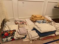 Overview of assorted linens including towels, wash cloths, aprons, table cloths, and sheets in multiple stacks on floor