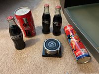 Collector items including four Coca-Cola glass bottles, two can coasters, cylindrical piggy bank, and black coaster set arranged on carpeted floor.