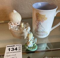 Photo of two Precious Moments figurines and one porcelain mug on glass shelf.
