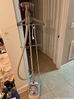 Full view of upright Pur garment steamer showing hanger, flexible steam hose, power cord, and water tank base on tile floor next to a white door.