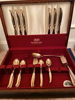 Set of flatware displayed in original red velvet-lined wooden case showing 6 knives, assorted forks, spoons, and serving utensils.